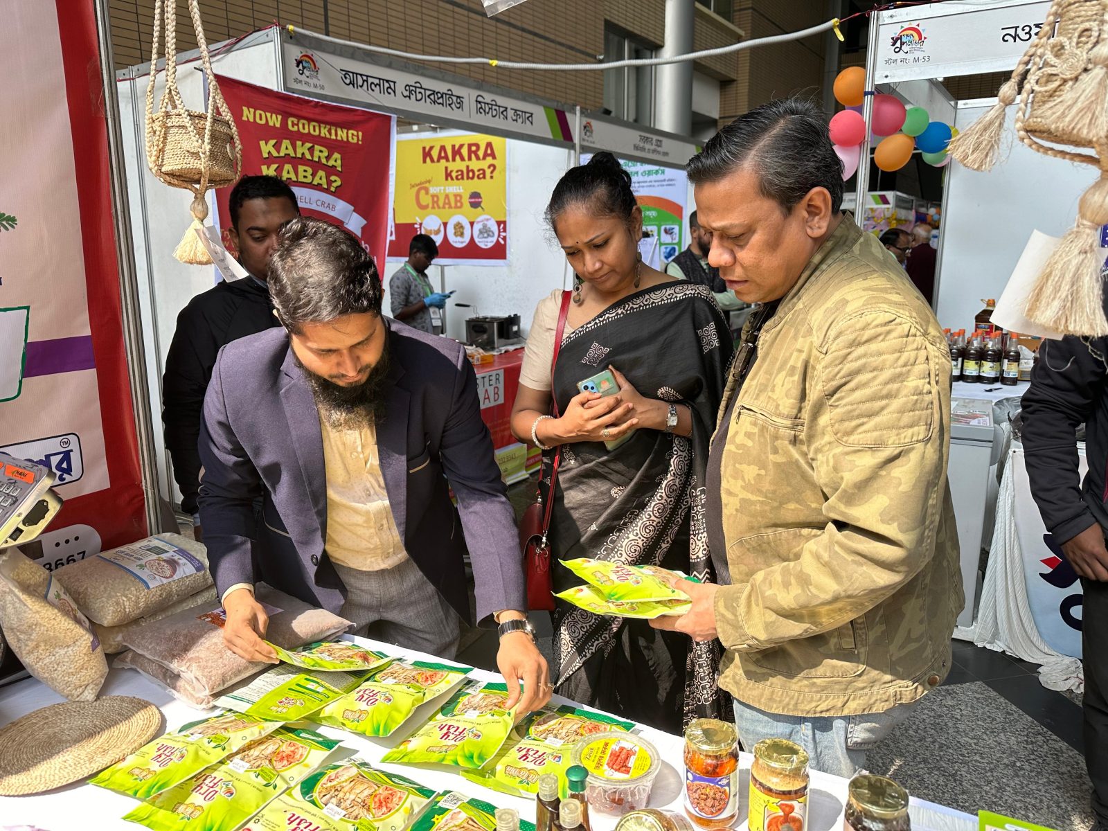 মেলার স্টলে ক্রেতারা লা লা দিয়ার শুঁটকি মাছ ও আচারের জার হাতে নিয়ে দেখছেন